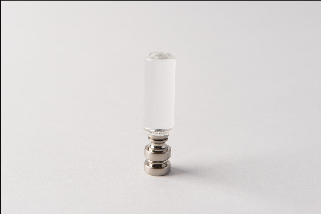 An Antique Brass Chinese Coin Finial stands upright on a plain white surface, centered in the image with a minimal background. The finial features a clear cylindrical glass body and a metallic antique brass base.